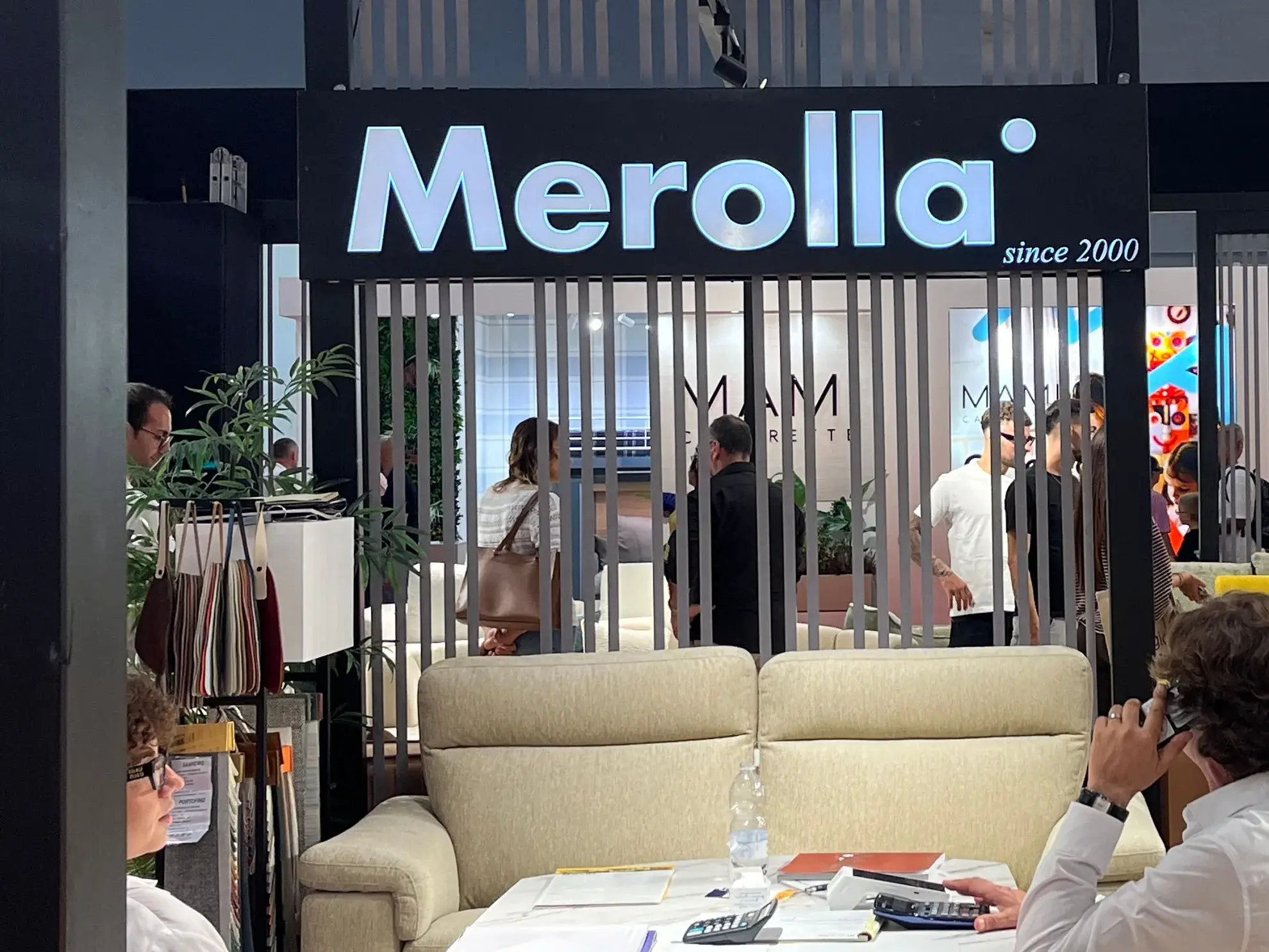 L’insegna Merolla sopra l’ingresso dello stand, con un divano color crema e il tavolo dove si sceglie il tessuto