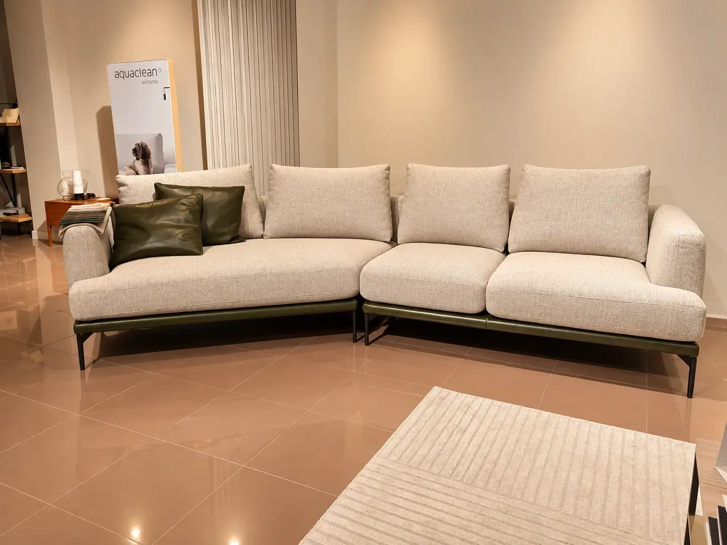 Divano angolare beige con cuscini verdi nello showroom, accanto a un tappeto chiaro