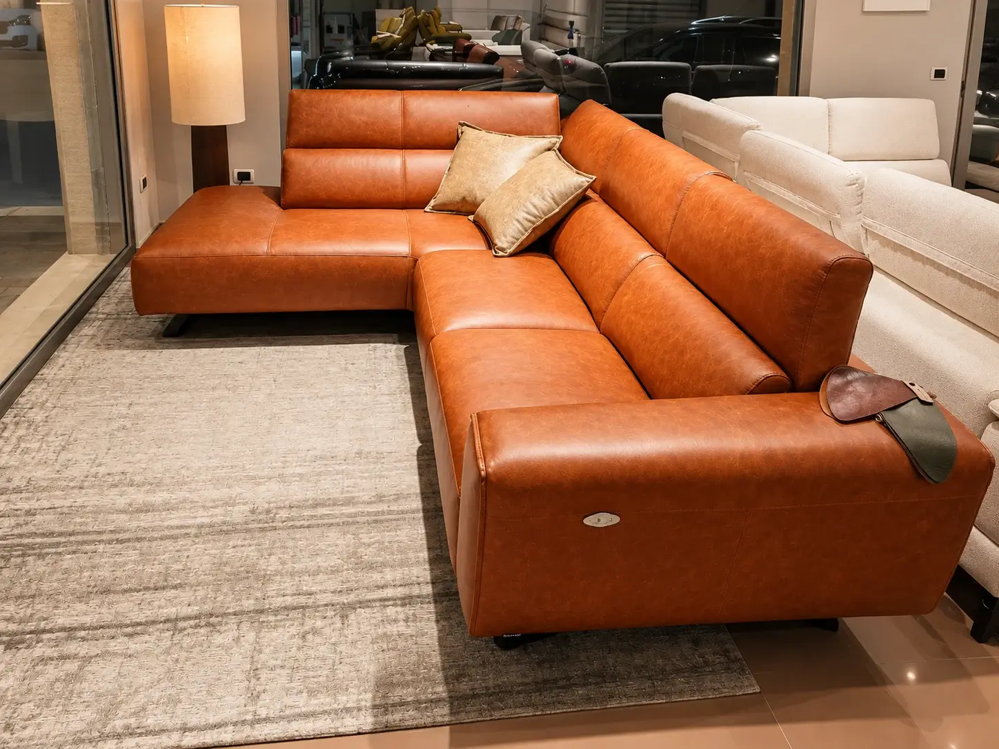 Divano angolare in pelle color cognac con cuscini chiari, su un tappeto grigio nello showroom